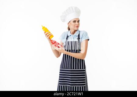 Jeune femme chef surprise avec tablier rayé et chapeau blanc regardant avec soin dans l'appareil photo avec des bouteilles de ketchup et de moutarde sur fond blanc Banque D'Images