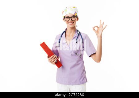 Jeune médecin souriant en uniforme et lunettes avec phonendoscope sur le cou, montrant heureusement geste ok regardant dans l'appareil photo avec dossier dans la main sur blanc Banque D'Images