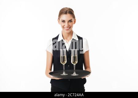 Jeune serveuse joyeuse dans un plateau de maintien uniforme avec des verres regardant avec plaisir dans l'appareil photo sur fond blanc isolé Banque D'Images