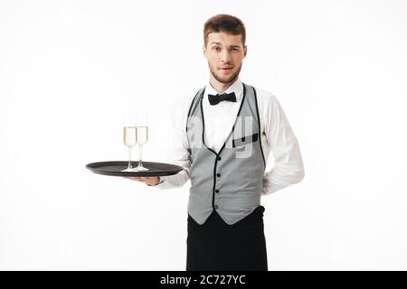 Jeune serveur dans un plateau de maintien uniforme avec des verres de champagne regardant avec soin dans l'appareil photo sur fond blanc Banque D'Images