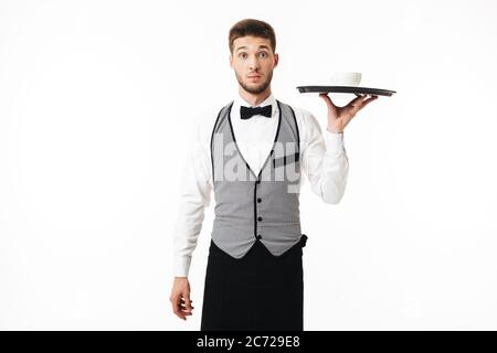 Jeune serveur choqué dans un plateau de maintien uniforme avec une tasse de café, regardant avec surprise dans l'appareil photo sur fond blanc Banque D'Images