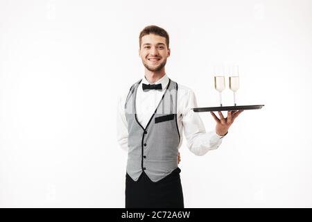 Joyeux serveur en uniforme regardant avec plaisir dans le plateau de maintien de l'appareil photo avec des verres sur fond blanc Banque D'Images