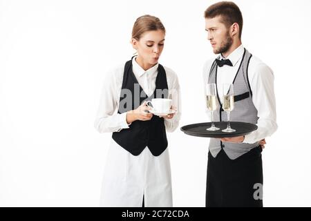 Une jeune serveuse soufflait sur une tasse de café tandis qu'un serveur sérieux tenait un plateau avec des verres de champagne bien pensé sur fond blanc Banque D'Images