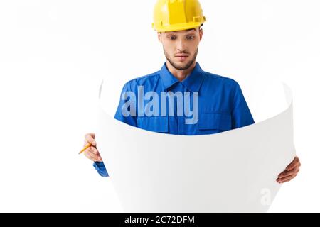 Jeune contremaître attentif en uniforme bleu et casque jaune regardant avec étonnement le plan de nouveau projet sur fond blanc Banque D'Images