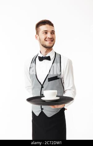 Jeune serveur joyeux dans un plateau de maintien uniforme avec une tasse de café à la main, regardant avec plaisir sur fond blanc Banque D'Images