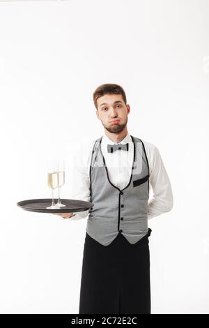 Jeune serveur dans un plateau de maintien uniforme avec des verres de champagne tiredly regardant dans l'appareil photo sur fond blanc Banque D'Images