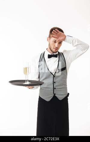 Jeune serveur fatigué dans un plateau de maintien uniforme avec des verres de champagne regardant soigneusement de côté sur fond blanc Banque D'Images