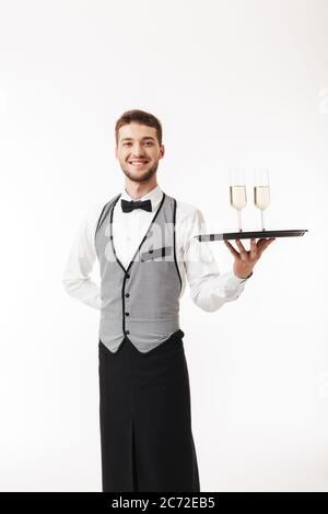 Jeune serveur gai en uniforme regardant avec joie dans le plateau de maintien de l'appareil photo avec des verres sur fond blanc Banque D'Images