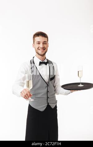 Jeune serveur souriant en uniforme, avec plateau de rangement plein de joie, offrant une coupe de champagne sur un fond blanc Banque D'Images