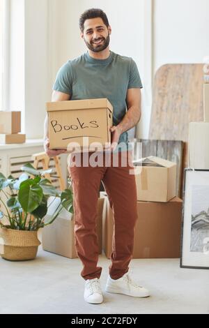 Portrait d'un jeune homme barbu tenant une boîte en carton avec des livres et souriant à l'appareil photo tout en se tenant debout dans son appartement Banque D'Images