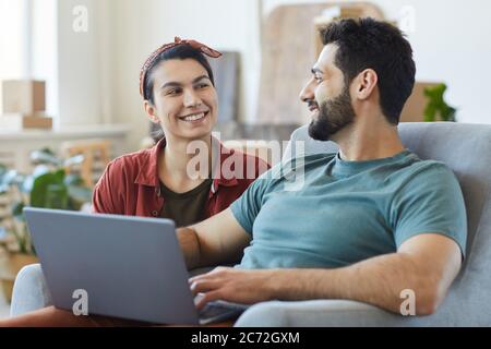 Jeune heureux couple assis sur un canapé parlant entre eux et utilisant un ordinateur portable tout en se reposant à la maison Banque D'Images