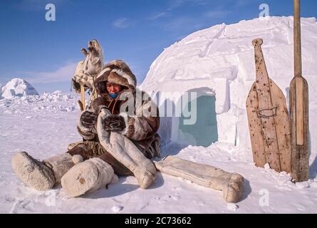 Une femme inspecte les doublures de bottes cousues à la main faites de peau de caribou tout en étant assise à l'extérieur de l'igloo. Elle est entourée d'outils de survie traditionnels. Banque D'Images