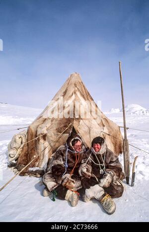 Homme et femme assis à l'extérieur de la tente de peau dans le haut arctique. Les outils traditionnels à côté d'eux et igloo peuvent être vus en arrière-plan. Banque D'Images