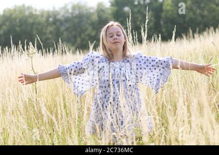 Une jeune fille assez hippie avec des cheveux blonds avec des bras étirés dans la longue herbe d'un champ de pâturage en été. Banque D'Images