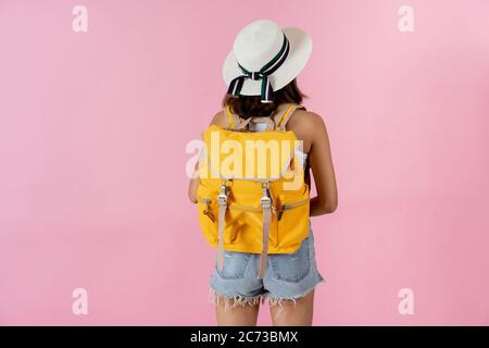 Vue arrière de la jeune femme en vacances portant un chapeau de soleil et portant un sac à dos jaune sur fond rose Uni Banque D'Images