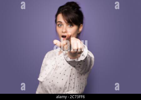 Choqué caucasien brune femme pointant vers la caméra avec la bouche ouverte sur un mur de studio violet Banque D'Images