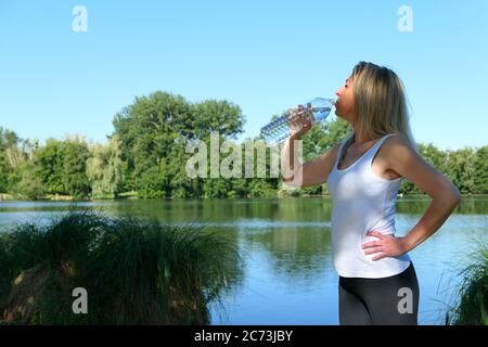 Femme blonde qui boit de l'eau de la bouteille pendant une séance de remise en forme dans la nature en été. Bien-être et style de vie sain. Banque D'Images