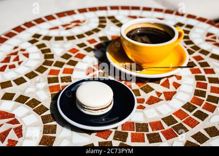 Macaron vanille au chocolat sur une assiette, une tasse jaune de café noir, sur une table en mosaïque de pierre d'un café par une journée ensoleillée Banque D'Images