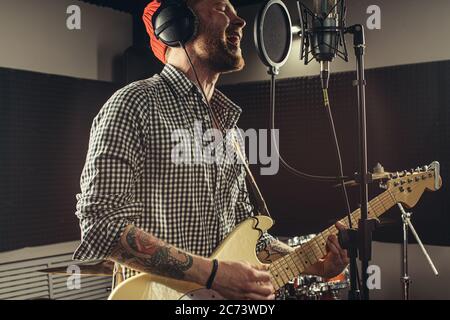 un jeune guitariste caucasien joue de la guitare et chante une chanson dans un studio d'enregistrement. un gars élégant de red hat chante émotionnellement une chanson, qui aime mu Banque D'Images