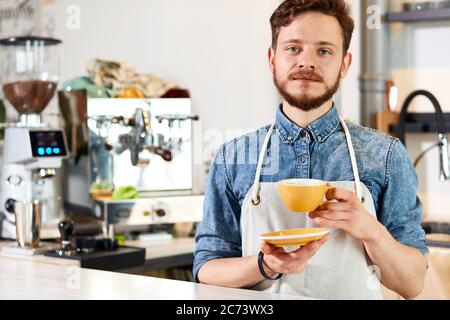 Le jeune barista professionnel se tient avec une coupe de café jaune, regarde l'appareil photo avec un visage calme, montre reconnaissant, prêt à donner du latte frais préparé Banque D'Images