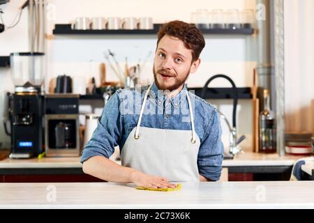 barista porte un jean et un tablier soigné, se tient sur un comptoir en bois, garde une main derrière le dos, met la main sur la table, nettoie avec un plumeau, regarde l'appareil photo, Banque D'Images