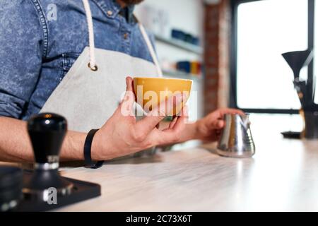 les mains d'un barman professionnel tiennent une petite tasse de café frais chaud avec les doigts, touchez le pot à lait en métal posé sur la table, prêt à servir le chapeau aromatique Banque D'Images