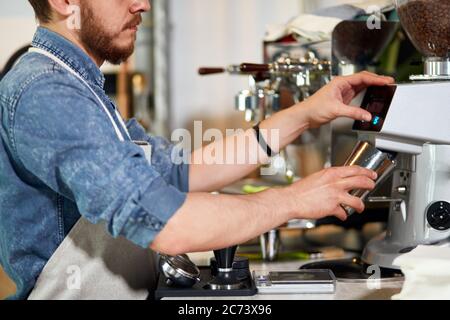 Les mains douces de la barbe le jeune barista tient la cuve en métal, le lait cuit à la vapeur, verse le café moulu, appuie sur le bouton de la machine à café dans le bar, professionnel c Banque D'Images