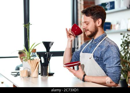 le barista optimiste et gai ferme les yeux avec plaisir, boit un cappuccino savoureux à l'aide d'un magnifique café rouge, aime le temps libre au travail dans un cadre moderne Banque D'Images
