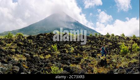 Vue panoramique sur le volcan arenal dans un parc national du Costa rica avec quelques nuages. Une femme marche en premier plan sur des rochers de lave avec Banque D'Images