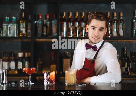 Portrait d'un beau jeune barman souriant, habillé en uniforme avec noeud papillon, sympathique souriant à l'appareil photo tout en se tenant au comptoir de bar avec des étagères o Banque D'Images