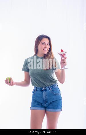 Belle jeune femme tenant un verre de martini avec des fraises et de la crème fouettée et une pomme verte tout en souriant à l'appareil photo sur un fond clair. Banque D'Images