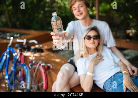 Photo en gros plan de l'homme tenant une bouteille d'eau dans le parc avec deux vélos à proximité. Jeune couple assis sur un banc et regardant avec plaisir dans la caméra pendant Banque D'Images