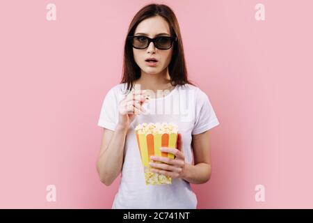 Jeune fille aux cheveux foncés, portant des lunettes 3d et mangeant du pop-corn dans un seau à rayures tout en regardant un film fascinant. Isolé sur le backgroun rose studio Banque D'Images