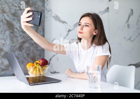 Jeune femme agitant la main et prenant photo de selfie sur téléphone portable à la cuisine blanche Banque D'Images