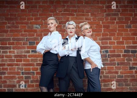 Trois femmes d'affaires blanches de race blanche adultes en costume de bureau posent près du mur de briques brunes. Deux d'entre eux ont croisé des bras, l'un d'eux tient la main dans la poche Banque D'Images