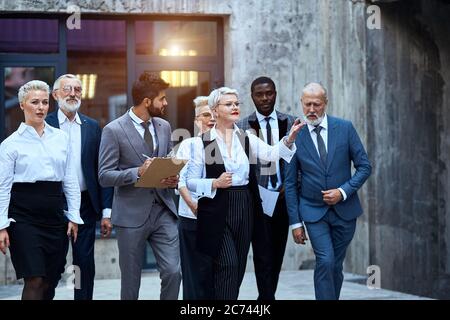 Groupe de sept hommes d'affaires qui se promère dans la rue. Blonde adulte caucasienne femme dire, homme en jaket gris écrire avis sur paper Banque D'Images