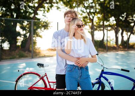 Jeune couple debout avec des vélos rouges et bleus à côté. Garçon debout avec une belle fille dans des lunettes de soleil et embrassant tout en passant du temps Banque D'Images