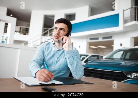 Un employé de concession automobile pensif parle au téléphone et écrit Banque D'Images