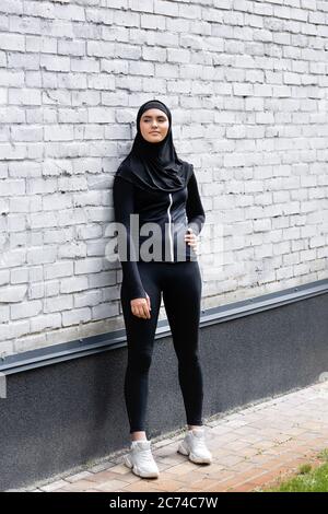 femme musulmane attirante dans le hijab debout avec la main sur la hanche près du mur de brique Banque D'Images