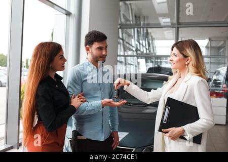 Une vendeuse de voitures dans un concessionnaire de voitures donne les clés à un jeune couple. Remise des clés de voiture aux nouveaux propriétaires Banque D'Images