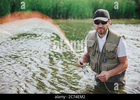 Jeunes pêcheurs pêchant sur le lac ou la rivière. Un homme sérieux concentré en robe et lunettes de soleil regardant directement sur l'appareil photo et tenant la ligne de pêche dans les mains Banque D'Images