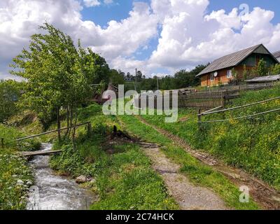 Paysage rural d'été avec un petit ruisseau de montagne tordant le long de la route, vieux pont fait main à travers la rivière, maison de campagne avec clôture en bois. Banque D'Images