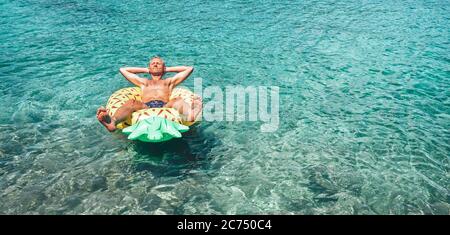 Homme se détendant en nageant sur un anneau gonflable de piscine d'ananas dans l'eau de mer cristalline. Image de concept de vacances imprudente. Banque D'Images