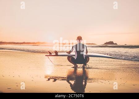 Surfeur avec surf à long bord assis sur la plage de sable de l'océan et en appréciant le ciel de coucher du soleil Banque D'Images