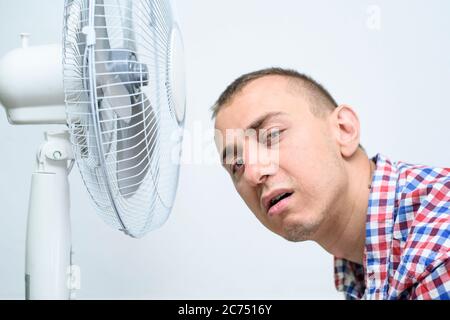 L'homme avec un chaume sur son visage souffre de la chaleur et essayant de refroidir près du ventilateur. Banque D'Images