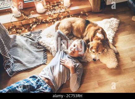 Garçon allongé sur le sol et souriant près de glisser son chien de beagle sur peau de mouton dans une ambiance chaleureuse. Moments paisibles de l'image de concept de maison confortable. Banque D'Images