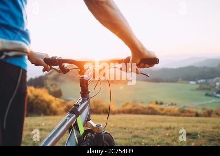 Homme à vélo reste au sommet d'une colline et profite du coucher du soleil. Homme mains sur le volant de vélo image de gros plan Banque D'Images