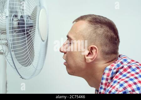 L'homme avec un chaume sur son visage souffre de la chaleur et essayant de refroidir près du ventilateur. Banque D'Images