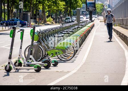 Un homme conduisant un scooter électrique sur une piste cyclable après une rangée de vélos partagés Velib garés à une station avec des scooters électriques Lime à Paris. Banque D'Images