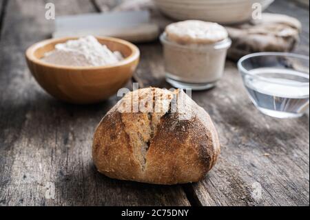 Pain au levain maison fraîchement cuit à côté des ingrédients - farine, eau et levure de base. Sur des panneaux de bois rustiques. Banque D'Images